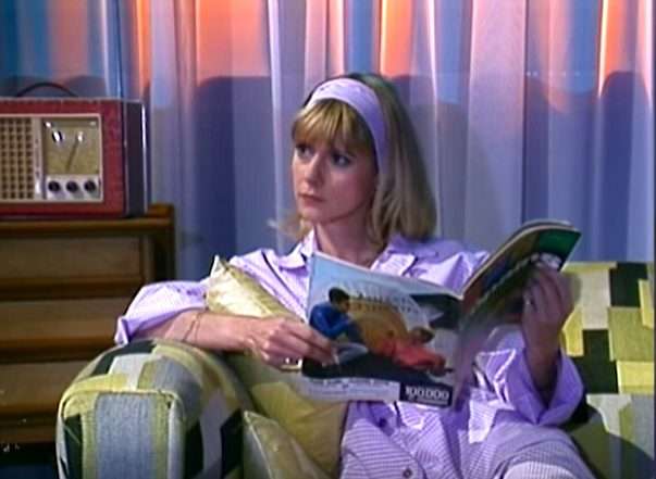 Dorothée lit un magazine assise dans un fauteuil, en chemise de nuit.